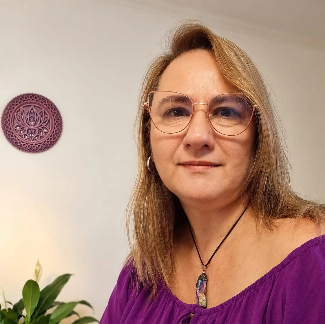 Rosi sorrindo levemente, sentada em frente a uma mandala de lótus na parede